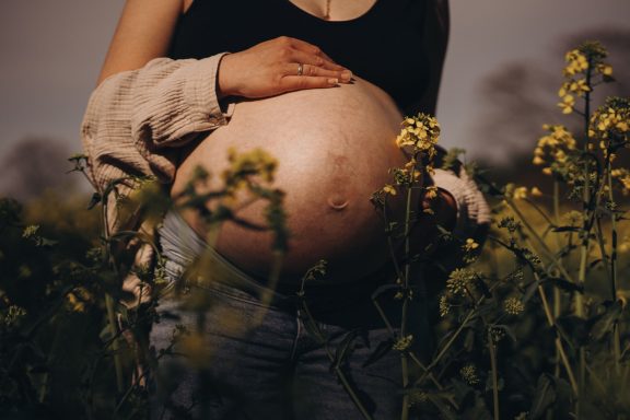 Schwangere Person mit freiem Bauch, die eine Hand auf den Bauch legt, umgeben von Blumen.