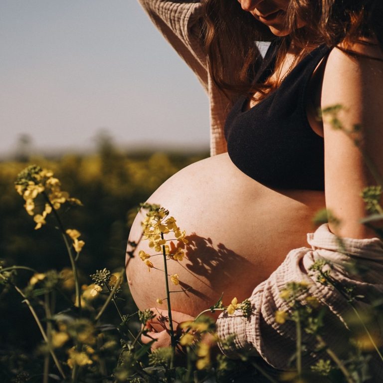 Schwangere Frau mit freiem Bauch, umgeben von gelben Blumen in der Natur.