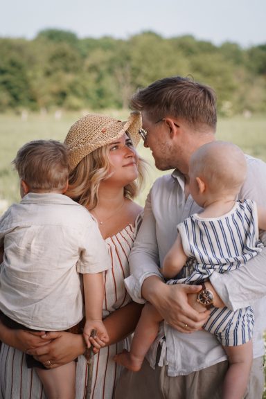 Familie mit zwei Kleinkindern steht eng zusammen, lächelt sich an und genießt die Zeit im Freien.